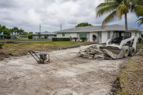 Driveway tear-out in progress at a 1970s Cape Coral block home, broken concrete slabs stacked beside a skid-steer and exposed white shell subgrade