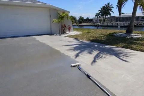 Half of a Cape Coral driveway darkened by newly applied clear sealer under midday sun, roller on an extension pole at the wet edge