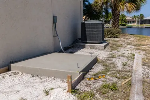 Fresh small concrete equipment pad with protruding anchor bolts beside a Cape Coral stucco wall, string line and level nearby