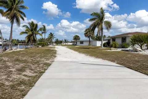 Finished pale concrete driveway in the foreground with a rutted white shell-gravel drive continuing toward a block home two lots down