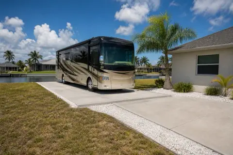 Wide concrete parking pad beside a Cape Coral home with a generic Class A motorhome parked on it, royal palm and canal beyond